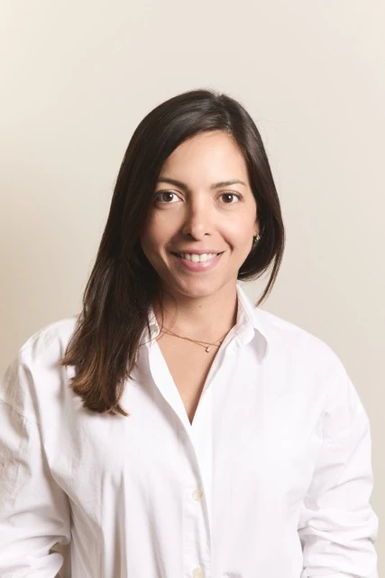 Retrato de Yasmin Morales sonriendo, con blazer crema y los brazos cruzados sobre fondo beige.