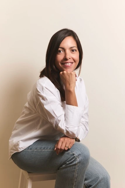 Yasmin Morales sentada en un taburete con camisa blanca y vaqueros, sonriendo con la mano bajo la barbilla.